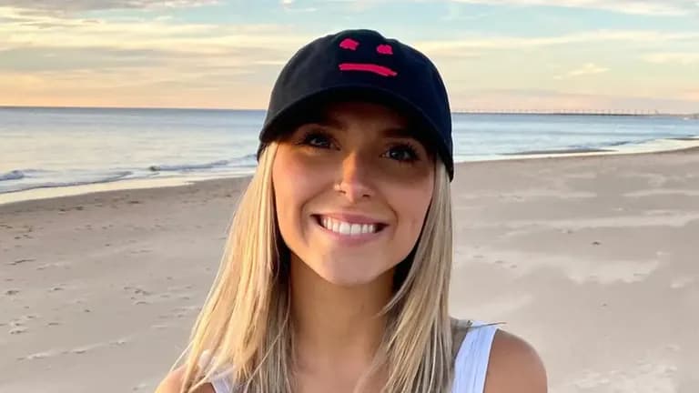 Emily Zugay smiling on a beach wearing a black cap with a red smiley face design, known for her viral logo redesigns on TikTok.