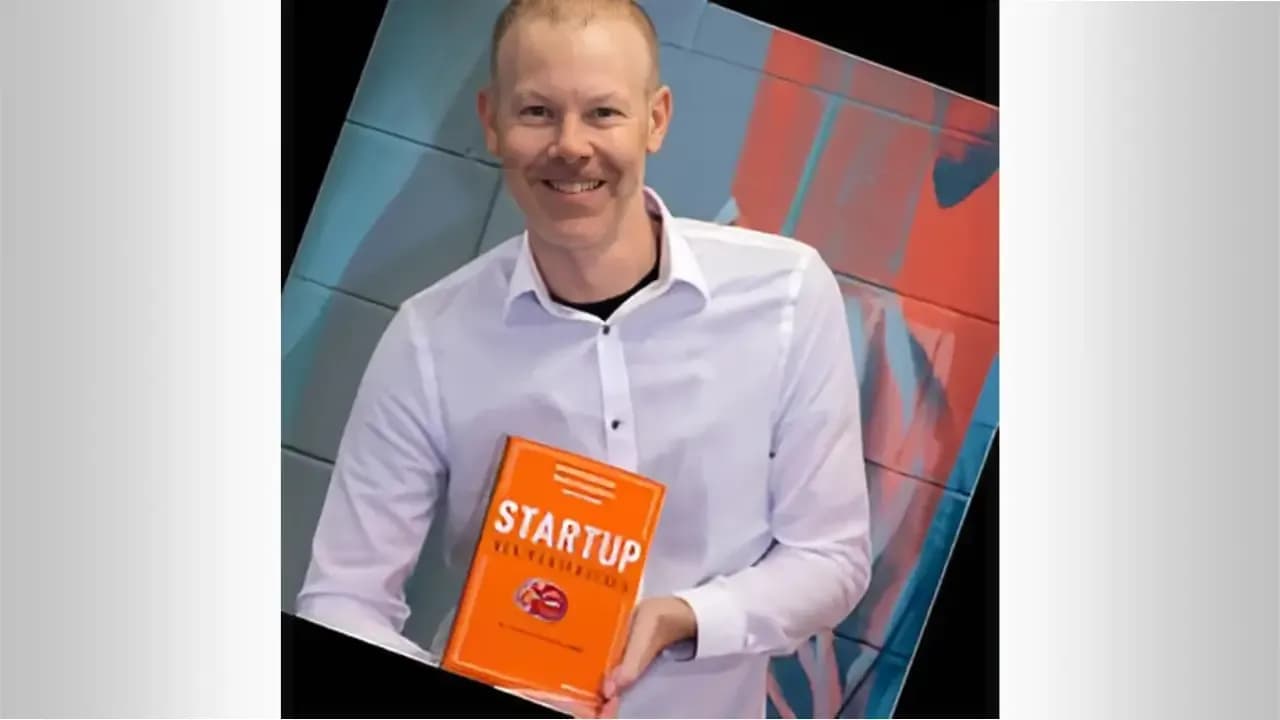 Steve Dunn, founder of Leapin Digital Keys, smiling and holding his orange “Startup Accelerators” book in front of a colorful tiled wall.