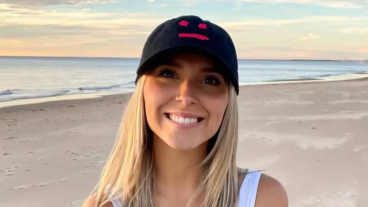 Emily Zugay smiling on a beach wearing a black cap with a red smiley face design, known for her viral logo redesigns on TikTok.