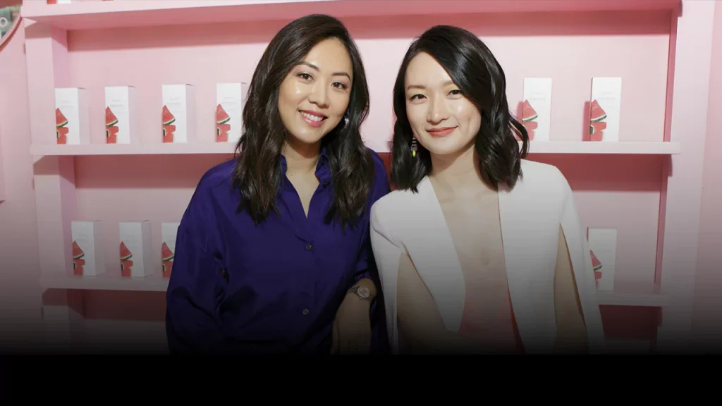 Glow Recipe founders Sarah Lee and Christine Chang posing in front of their signature skincare products in 2025