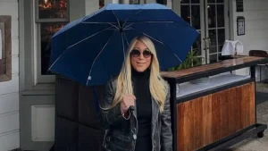 Lori Greiner holding a blue umbrella, wearing sunglasses and a leather jacket outside a restaurant.