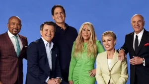 Mark Cuban stands with fellow Shark Tank judges Daymond John, Robert Herjavec, Lori Greiner, Barbara Corcoran, and Kevin O’Leary on a blue background.