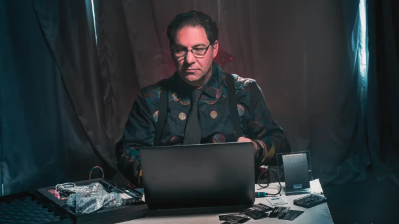 A serious man in a dark, patterned shirt sits at a desk with a laptop and hacking tools, representing Kevin Mitnick, once the world’s most wanted hacker.