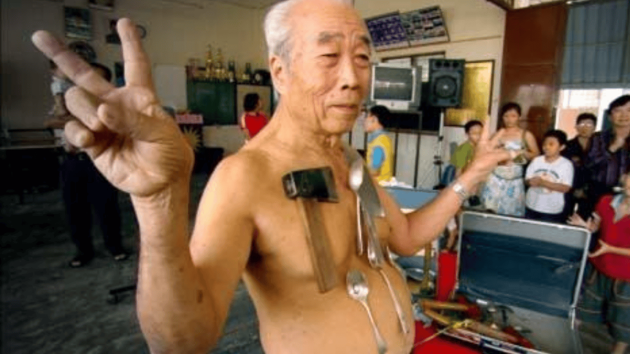 Elderly man known as the Human Magnet standing shirtless with metal objects like spoons and hammers sticking to his chest, surrounded by spectators.